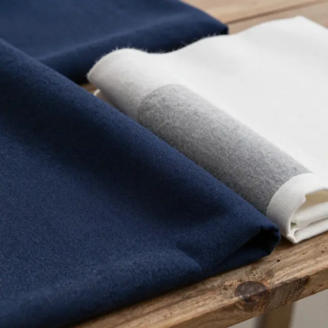 Close-up photograph of premium fabrics in shades of dark blue, gray, and white laid out on a wooden table in a bright studio, showing texture details that suggest insulation and windproof properties, evoking warmth and functionality for Antarctic conditions.